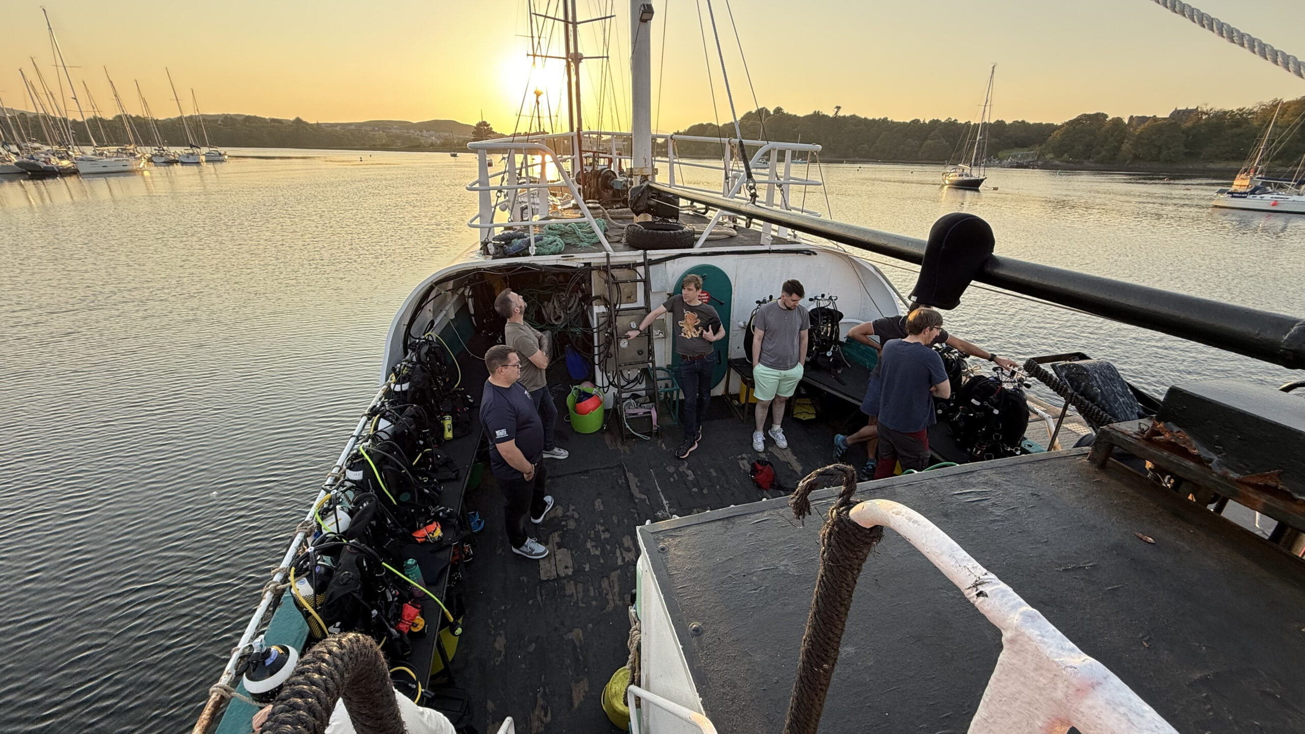 Six male divers stood on the dive deck of MV Honeydew with the sun setting behind.