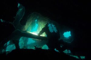 a diver wearing a drysuit and twinset looks through an opening in a wreck