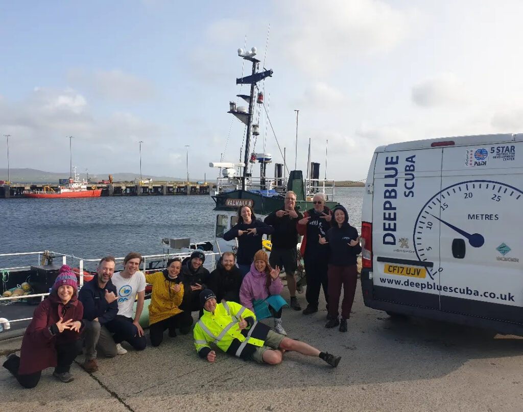 Group of 12 divers posing with MV Valhalla in background and white van with club logo